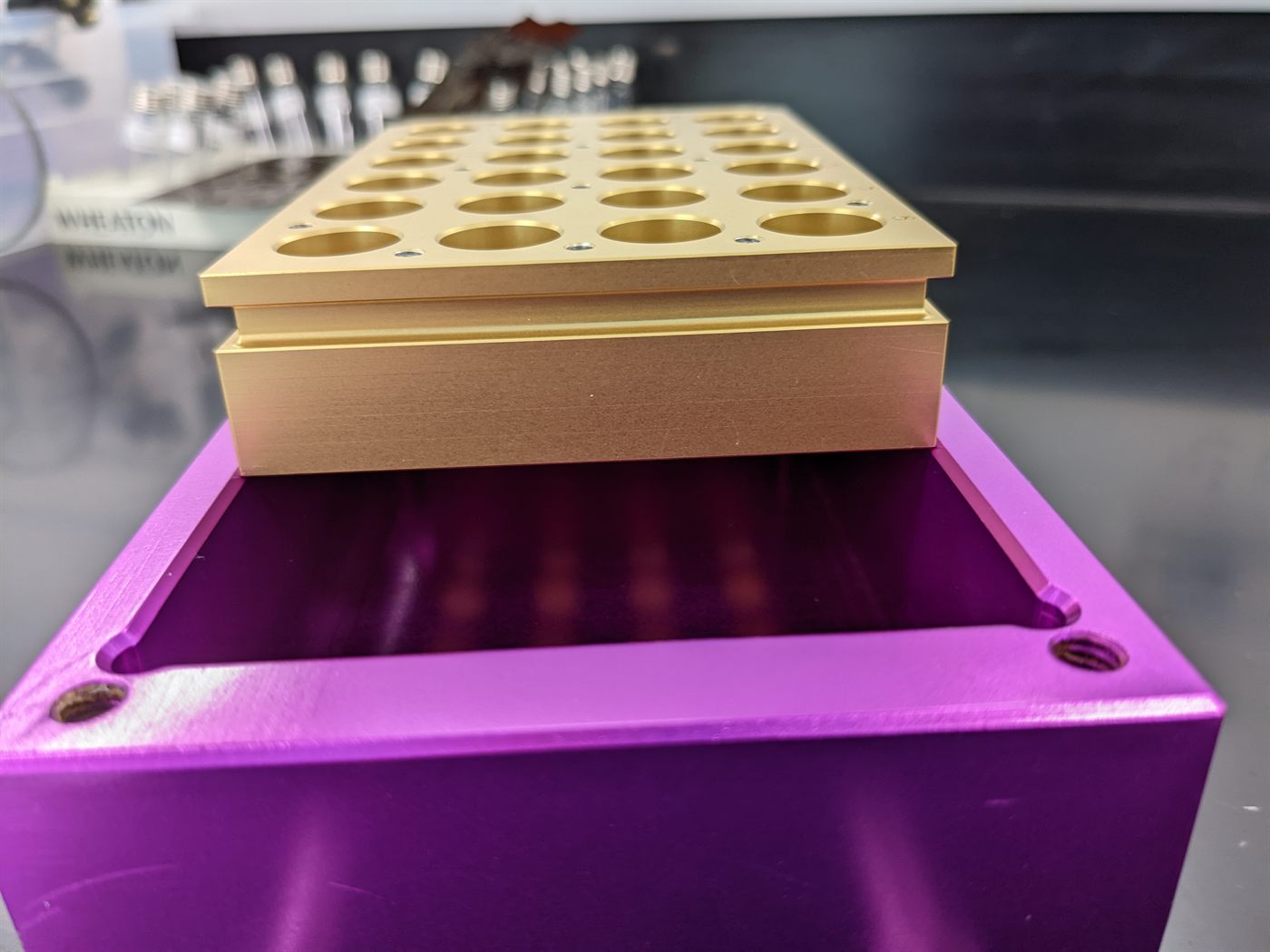 Gold anodized aluminum vial rack with 28 counterbored pockets, sitting on a magenta anodized aluminum tray